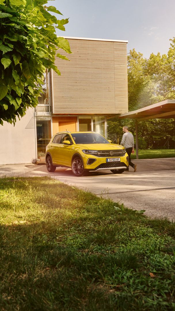 Ein gelber VW T-Cross steht in der Auffahrt eines modernen Wohnhauses mit großen Bäumen.