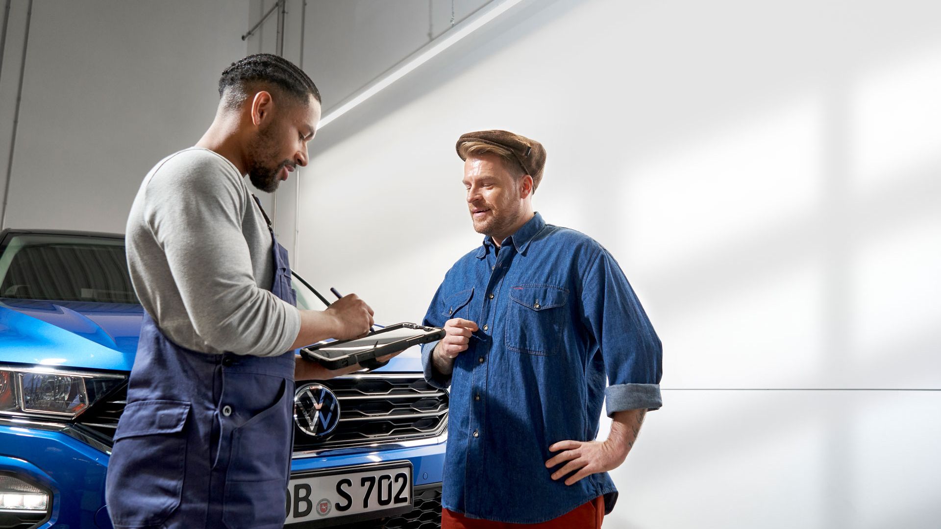 Zwei Personen stehen vor einem blauen Volkswagen und unterhalten sich. Die Person auf der linken Seite trägt eine Arbeitskleidung und hält einen Klemmbrett. Die Person auf der rechten Seite trägt legere Kleidung und hat eine Hand in die Hüfte gestützt.