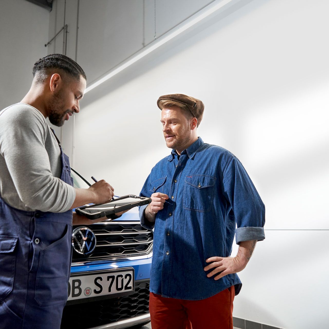 Zwei Personen stehen vor einem blauen Volkswagen und unterhalten sich. Die Person auf der linken Seite trägt eine Arbeitskleidung und hält einen Klemmbrett. Die Person auf der rechten Seite trägt legere Kleidung und hat eine Hand in die Hüfte gestützt.