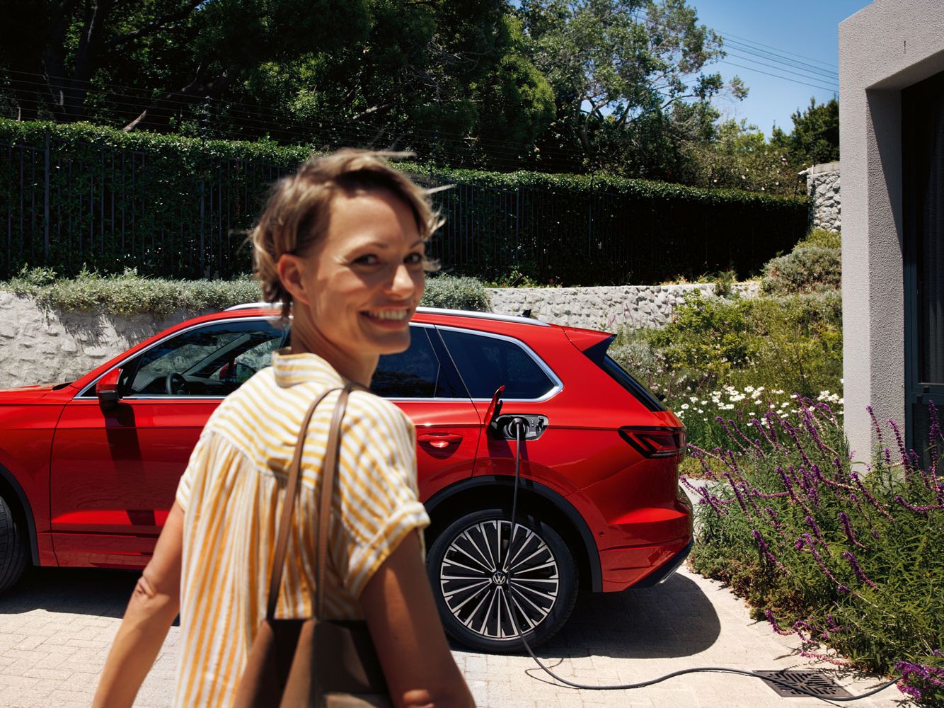 Im Vordergrund eine lächelnde Frau, im Hintergrund lädt ein VW Touareg Elegance eHybrid an einer Ladesäule.