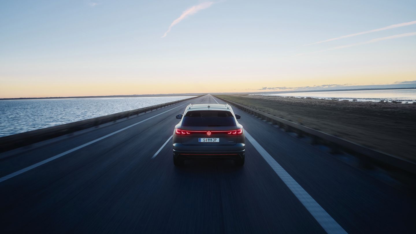 Ein Touareg R fährt auf einer leeren Straße entlang eines Gewässers bei Sonnenuntergang. Das Fahrzeug ist von hinten zu sehen. Die Straße erstreckt sich gerade in die Ferne.