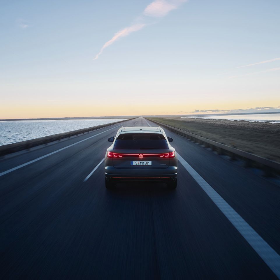 Ein Touareg R fährt auf einer leeren Straße entlang eines Gewässers bei Sonnenuntergang. Das Fahrzeug ist von hinten zu sehen. Die Straße erstreckt sich gerade in die Ferne.