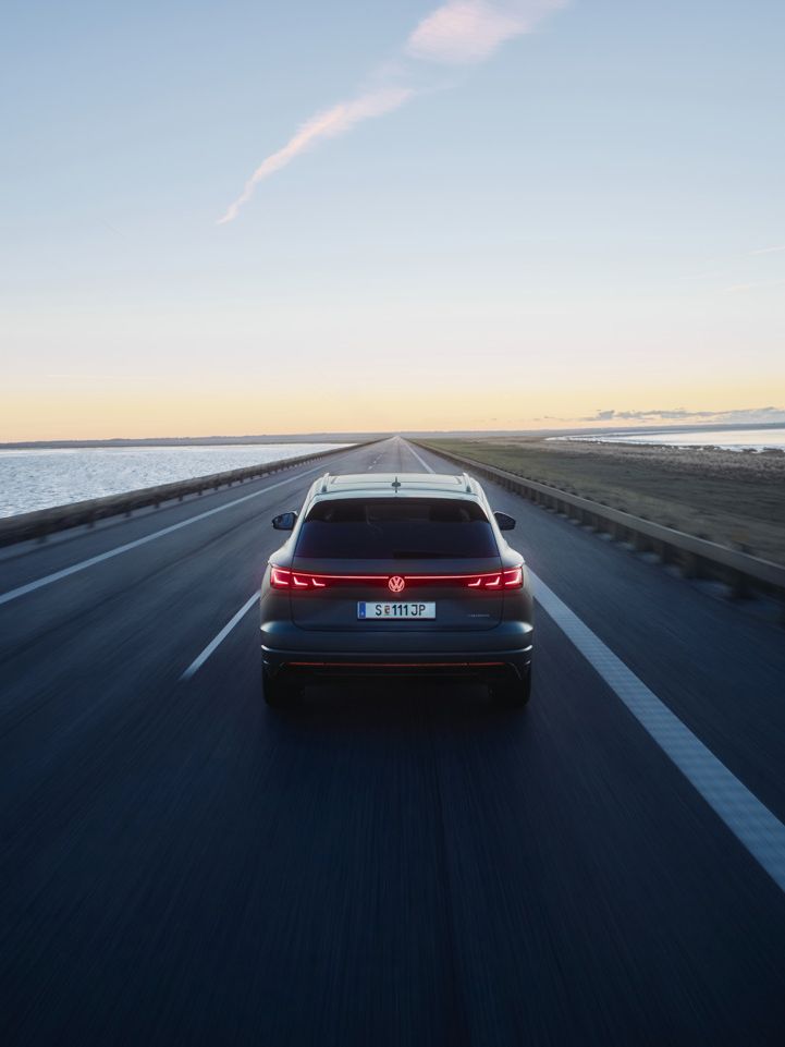 Ein Touareg R fährt auf einer leeren Straße entlang eines Gewässers bei Sonnenuntergang. Das Fahrzeug ist von hinten zu sehen. Die Straße erstreckt sich gerade in die Ferne.