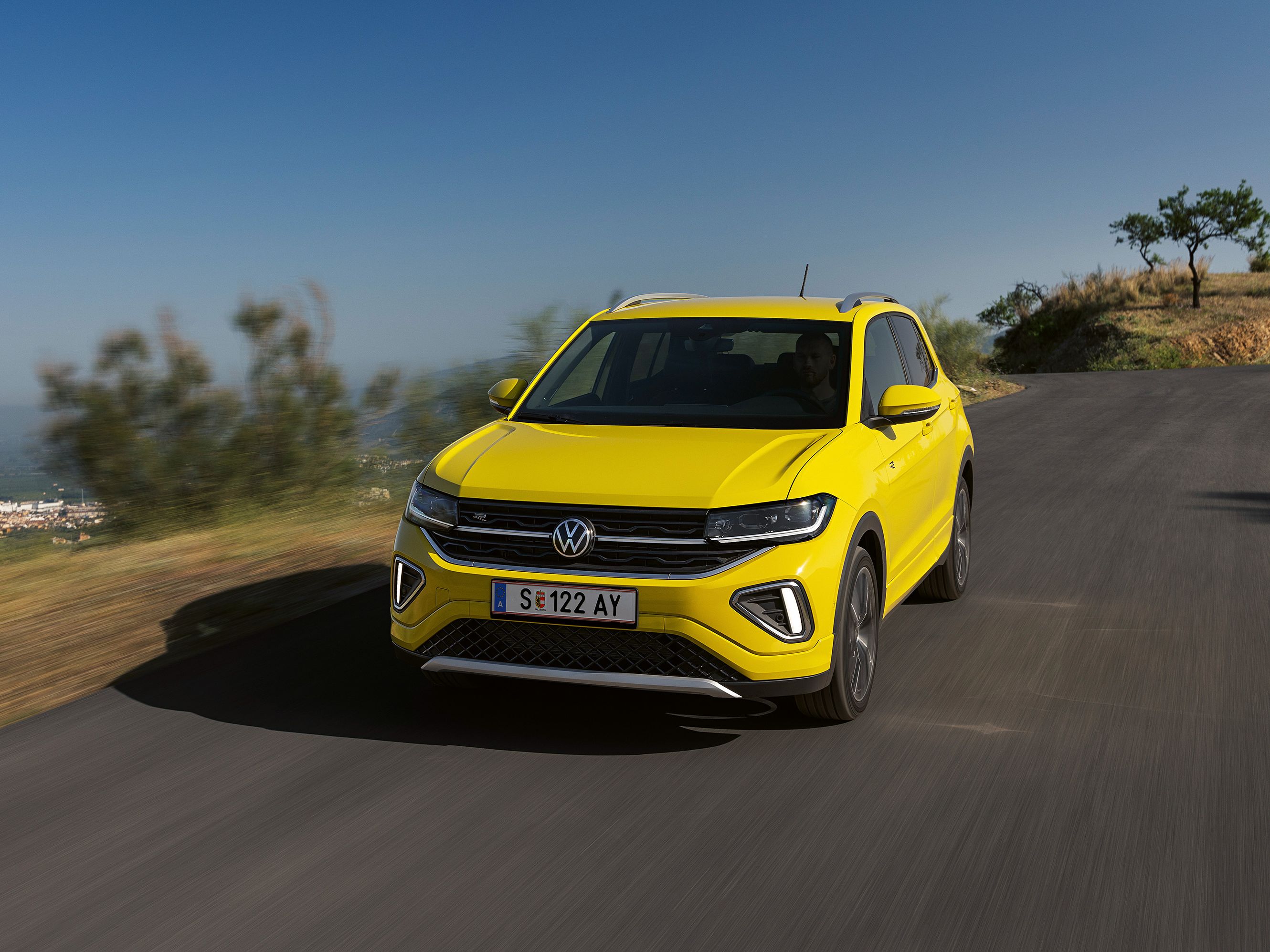 Gelber Volkswagen SUV fährt auf einer Straße in einer ländlichen Gegend. Hintergrund zeigt verschwommene Natur und stark bewölkten Himmel. Das Fahrzeug ist frontal zu sehen.